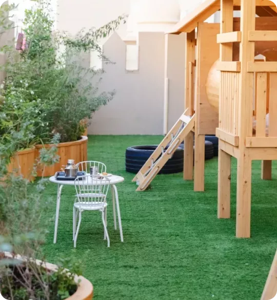 Outdoor nursery play area at Blossom Nursery & Preschool with a wooden climbing frame, slide, and small table set on green artificial grass.